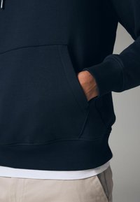 Sudadera azul marino con un bolsillo frontal, puños y dobladillo acanalados. La tela parece suave y ligeramente texturada, superpuesta a una camisa blanca.