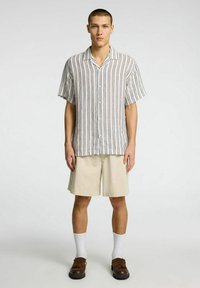 Camisa de botones de manga corta con patrón de rayas verticales en tonos claros. Combinada con pantalones cortos beige y zapatos tipo slip-on marrones, completando el conjunto.