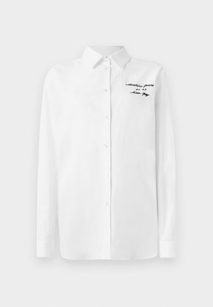 Camisa blanca de manga larga con botones y cuello, con texto bordado en negro "Moschino Jeans" en la parte superior izquierda del pecho.