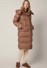 Brązowa kurtka puchowa o prostokątnym kroju, z kapturem, dużymi kieszeniami i gładką fakturą. Nosi na sobie jasnobeżowe spodnie i brązowe buty.