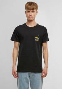 T-shirt en coton noir avec un graphique sur la poche avant représentant un bol de nouilles avec des baguettes, le texte "SEND NOODS" en jaune, design col rond.