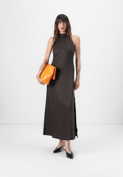 Mouwloze, donkerbruine maxi-jurk met een hoge hals en een zijschuif. Model houdt een oranje clutch vast; zwarte open teen schoenen maken de look af.