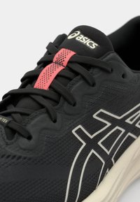 Chaussure de sport noire avec une tige texturée, logo blanc et accents, et un porte-lacets rose avec des perforations, alliant fonctionnalité et design.