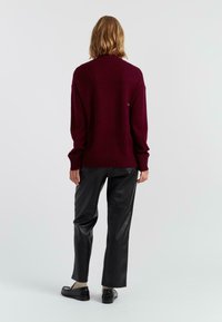 Bordeaux gestrickter Pullover mit gerippten Bündchen und Saum, kombiniert mit schwarzen Fake-Lederhosen, von hinten betrachtet. Einfaches und sauberes Design.