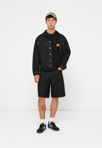 Black denim jacket with silver buttons, layered over a black t-shirt with an orange pocket label. Black denim shorts and black sneakers with white stripes.