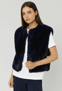 Gilet en fausse fourrure marine avec un col rond, sans manches et des fermetures à pression sur le devant, porté sur un haut blanc. Texture douce, design simple.