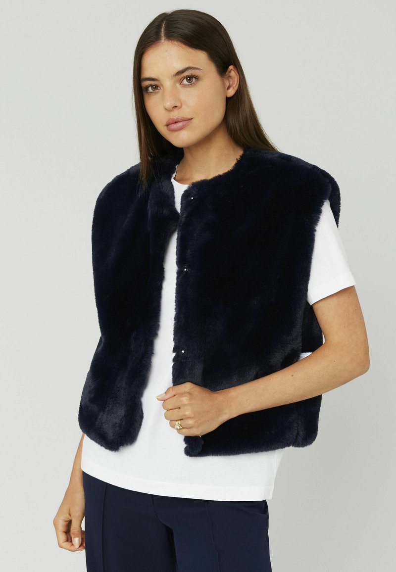 Gilet en fausse fourrure marine avec un col rond, sans manches et des fermetures à pression sur le devant, porté sur un haut blanc. Texture douce, design simple.