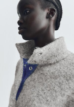 Siv gray pulover z visokim ovratnikom, ki ima modri trikotni detajl in srebrne pritiske. Mehka tekstura s rahlo puhasto podobo.