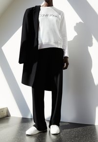 Felpa bianca con logo "Calvin Klein", cappotto nero appoggiato sulla spalla, pantaloni neri larghi e scarpe bianche slip-on. Texture lisce.