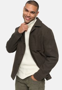 Chaqueta de lana marrón con cierre de botones, bolsillo en el pecho, llevada sobre un cuello alto de punto blanco. Los pantalones en color verde oliva completan el conjunto.