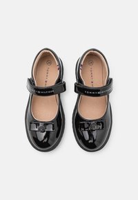 Zapatos de charol negro con puntera redonda, cierre con correa y detalle de lazo. Presentan un logo en relieve y un forro interior texturizado. Talla 28.