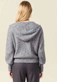 Grå stickad tröja med huva, med ribbat mönster och avslappnad passform. Långa ärmar och en åtsittande midja fulländar designen.