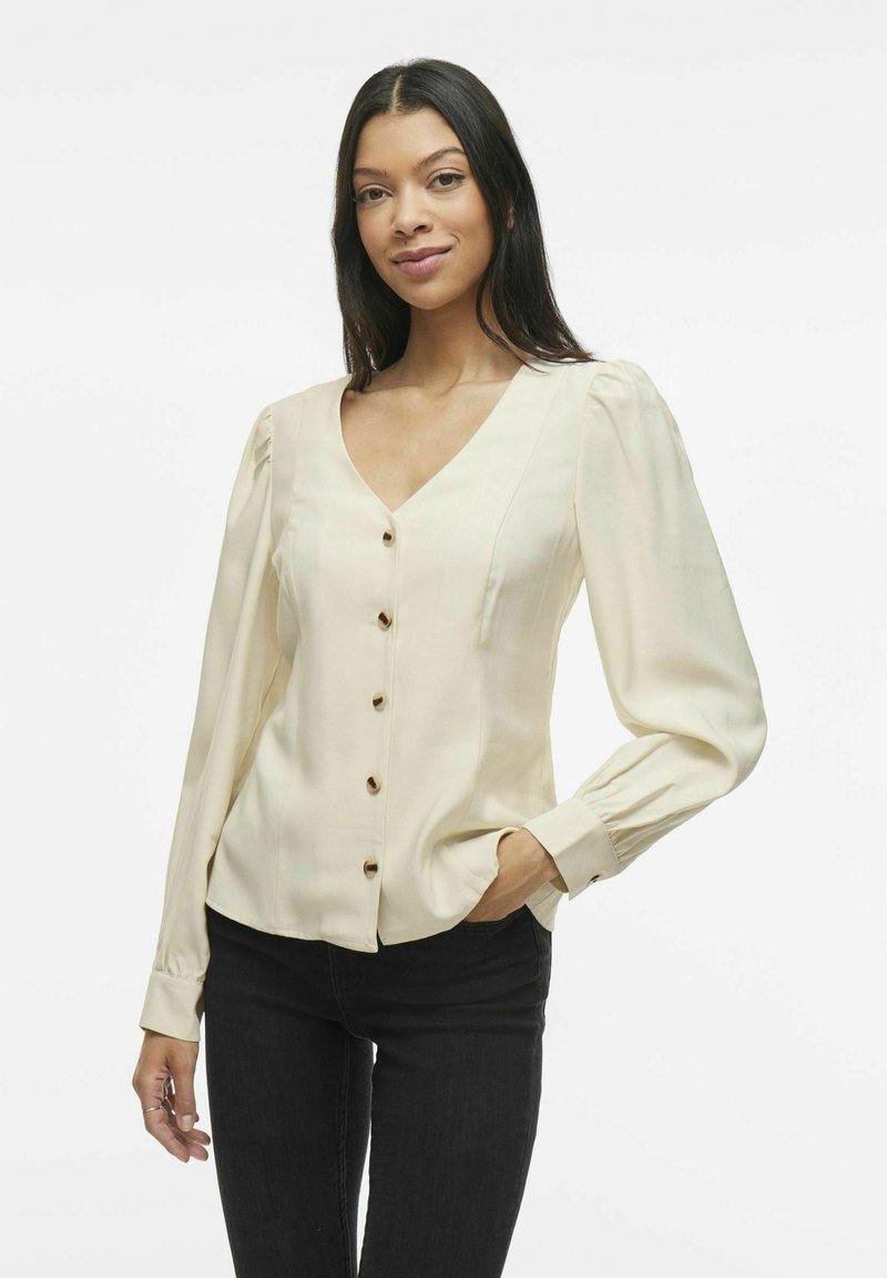 Blusa color crema con scollo a V, maniche lunghe a palloncino, chiusura finta con bottoni e texture liscia. Abbinata a jeans scuri.