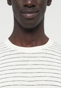 Άσπρο πλεκτό πουλόβερ με λεπτές γκρίζες οριζόντιες ρίγες, στρογγυλή λαιμόκοψη και απαλή υφή. Κοντινή όψη γύρω από το λαιμό και το πρόσωπο.