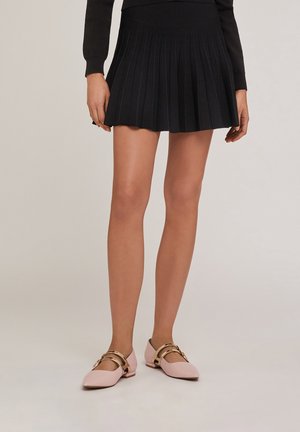 Chaussures plates roses avec des accents de boucles dorées, assorties à une jupe plissée noire. La tenue présente un design simple et élégant.