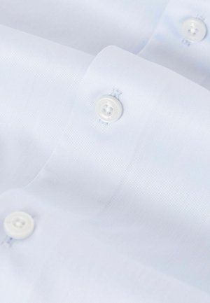 Lähedal võetud foto helesinisest dressipluusist, kus on näha valged nööbid ja tekstureeritud kangas diagonaalsete kootud mustritega.