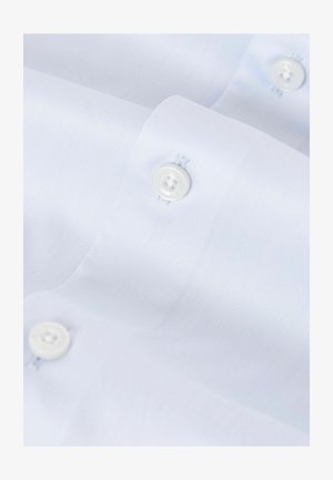 Lähedal võetud foto helesinisest dressipluusist, kus on näha valged nööbid ja tekstureeritud kangas diagonaalsete kootud mustritega.