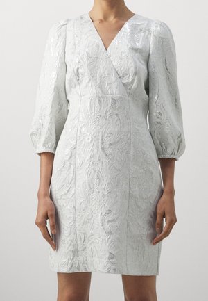 Kobieta ubrana w białą teksturowaną sukienkę kopertową z rękawami 3/4 typu puff oraz kwiatowym tłoczonym wzorem, stojąca na gładkim jasnym tle.