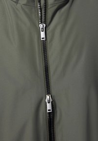 Olivegrüne Jacke mit einem auffälligen schwarzen Reißverschluss, glatter Textur und einem schlanken Design ohne zusätzliche Muster oder Akzente.