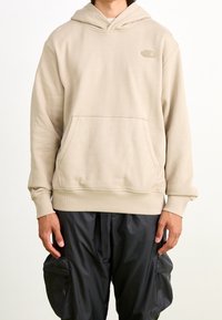 Béžová mikina s kapucí z měkké látky s klokaní kapsou a žebrovanými manžetami. Na hrudi je malé vyražené logo.