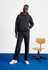 Juodas sweatshiertas su priekinėmis kišenėmis, derinamas su juodais kelnėmis ir baltais sportbačiais su raudonomis juostelėmis. Paprastos fono, ant medinės kėdės.