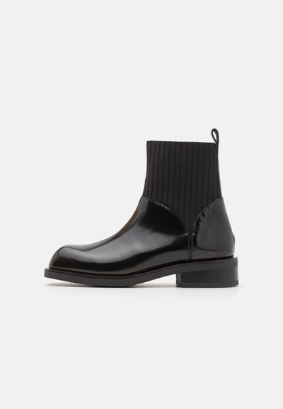 Botas de tornozelo pretas com acabamento em pele brilhante, parte superior em tecido preto canelado e salto baixo em bloco. Bico arredondado e laço na parte de trás.