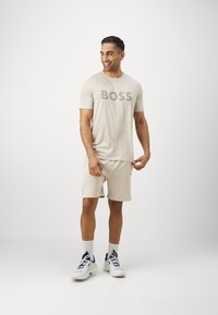 Camiseta beige con el logo "BOSS" en gris, combinada con pantalones cortos beige a juego. Medias blancas y zapatillas gris/negro. Diseño simple y atlético.
