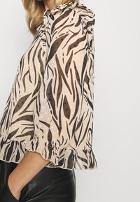 Transparent bluse med zebra-striber i brunt og beige, med flæseærmer og en struktureret overflade, kombineret med mørke bukser.