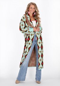 Lungo cardigan con motivi geometrici nei colori turchese, nero e rosso. Presenta dettagli in frange lungo il colletto. Indossato sopra una maglietta bianca e jeans svasati.