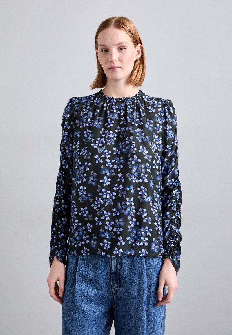 Schwarze Bluse mit floralem Muster in Blau, mit gerafftem Ausschnitt und gerafften Ärmeln. Getragen mit hellblauen, locker sitzenden Denim-Hosen.