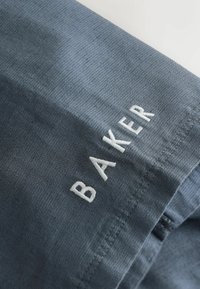 Tissu bleu texturé avec des lettres blanches brodées formant le mot « BAKER » le long du bord, montrant des points de couture détaillés.