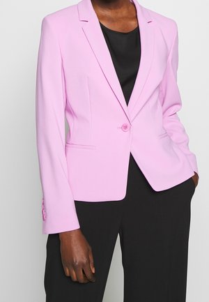 Blazer rose clair en tissu tissé, dotée de revers crantés, de fermeture à un bouton et de deux poches avant. Porté par-dessus un top noir.