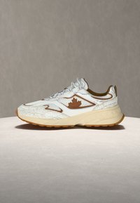 Baskets blanches et beiges avec logo feuille d'érable marron, design texturé et semelle épaisse, présentées sur une surface claire contre un fond uni.