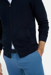 Marineblauer, mit Reißverschluss versehenen Cardigan aus strukturierter Stoff, mit Reißverschluss, Seitentasche und gerippten Bündchen, kombiniert mit hellblauen Hosen.