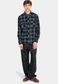 Groene en zwarte geruite flanellen shirt met twee borstzakken, knopen-sluiting, ronde kraag en lange mouwen, gecombineerd met donkere spijkerbroek.