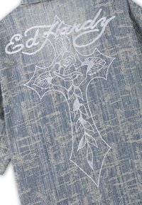 Dos de veste en denim avec croix blanche brodée et motif de rose, avec « Ed Hardy » inscrit au-dessus sur un tissu bleu texturé.