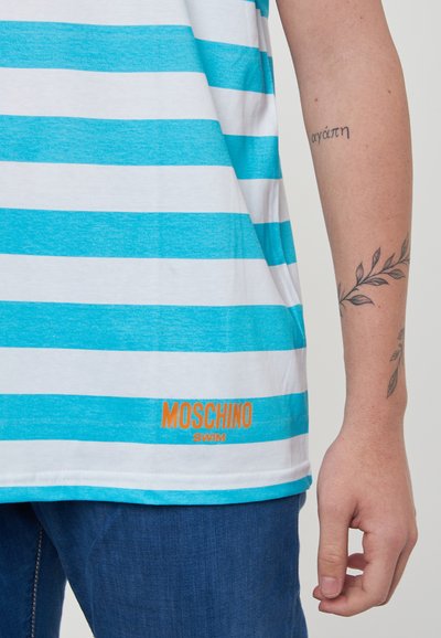 T-shirt a righe verticali in turchese e bianco, con il logo "Moschino Swim" in arancione. Tessuto morbido con una vestibilità comoda, indossato con jeans blu.
