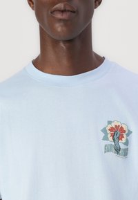 Camiseta de algodão azul-claro com um gráfico floral que apresenta uma mão e o texto "SANTA RUZ" em verde, localizado na área do peito esquerdo.