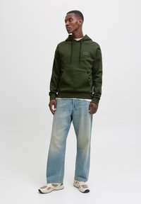 Green hoodie with drawstring, front pocket, and logo. Paired with light blue wide-leg jeans and beige sneakers with patterned accents.