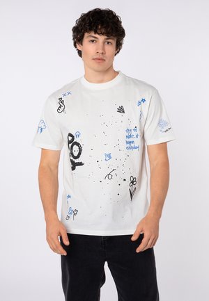 Junger Mann mit lockigem Haar trägt ein weißes T-Shirt mit blauen und schwarzen Kritzeleien und Text, kombiniert mit einer schwarzen Hose, stehend vor weißem Hintergrund.