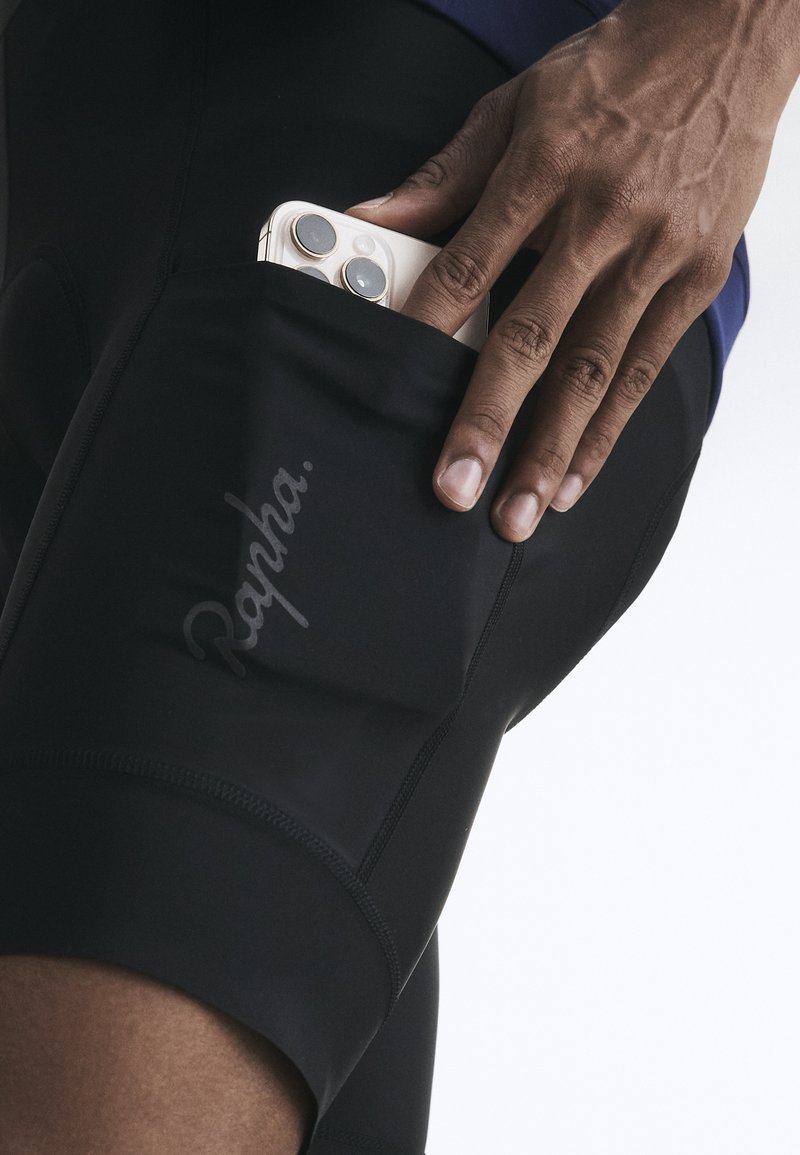 Hand schiebt ein weißes Smartphone in die Seitentasche einer schwarzen Rapha Radhose vor einfarbigem Hintergrund.