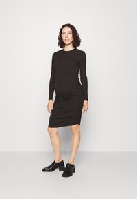 Robe noire à manches longues côtelées avec ruching sur les côtés, silhouette ajustée, longueur genou, assortie à des chaussures à talons noires. Texture lisse, design minimal.