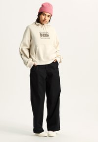 Beige hoodie met grafische print, voorkeurszak, zwarte wijde broek en witte schoenen. Roze gebreide beanie, casual en oversized fit.