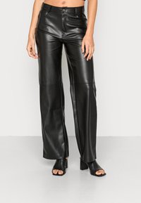 Pantalons large en cuir noir à taille haute, avec une fermeture à bouton sur le devant et une texture lisse. Portés avec des talons ouverts noirs.