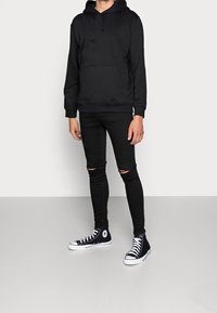 Sudadera negra con bolsillo canguro, combinada con jeans skinny negros rasgados y zapatillas altas negras. Tejido suave, ajuste casual.