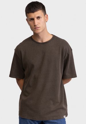 Νέος άνδρας με κοντά σκούρα μαλλιά, που φοράει μια φαρδιά καφέ κοντομάνικη μπλούζα και μπλε παντελόνι, στέκεται με τα χέρια πίσω από την πλάτη.