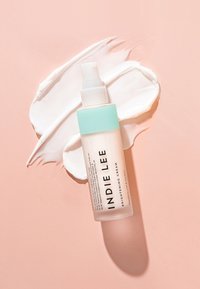 Flacon de crème éclaircissante avec un capuchon vert menthe, un corps transparent et un texte noir ; échantillon de produit blanc crémeux sur un fond rose doux.