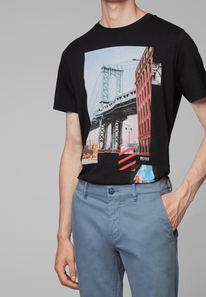 Personne portant un t-shirt noir avec un motif imprimé d'un pont de ville, un bâtiment en briques rouges et un petit détail de ciel bleu, associé à un pantalon bleu.