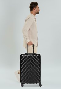 Mann in beiger Kleidung geht, während er einen großen schwarzen Hartschalenkoffer mit vier Rädern und einem ausziehbaren Griff hinter sich herzieht.
