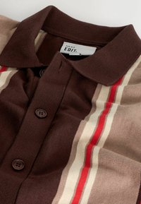 Braunes Strick-Polohemd mit Vorderknöpfen und senkrechten Streifen in Beige, Creme und Rot an den Seiten, beschriftet mit NEXT EDIT.
